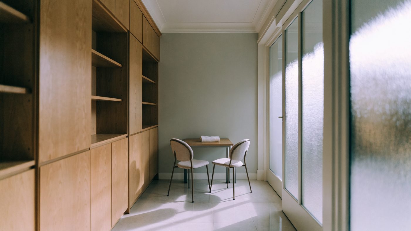 A calm private consulting room in a high-street pharmacy, pale wood cabinetry and a small desk with two chairs beside a frosted glass door