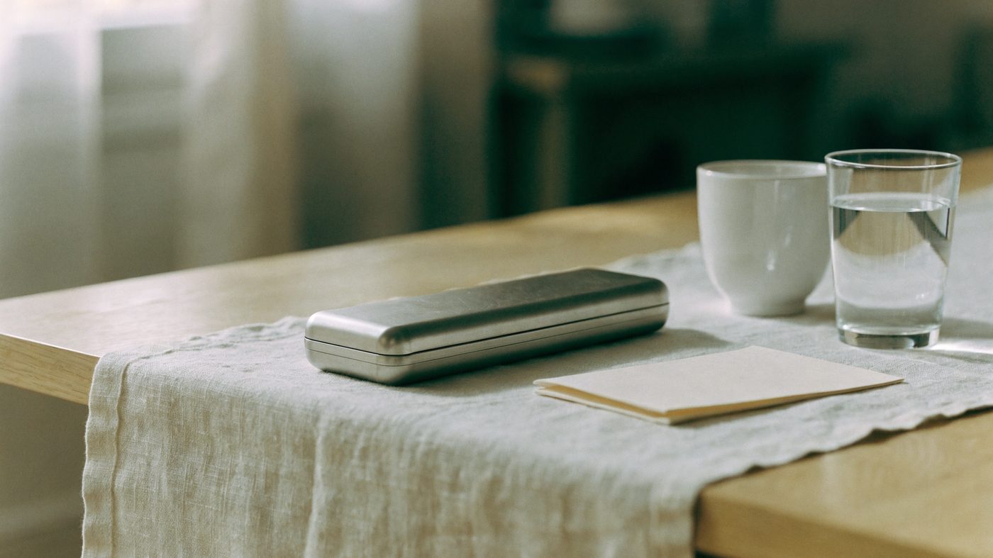 A pale oak table with a closed instrument case, a folded card, a cup and a glass of water on a linen runner