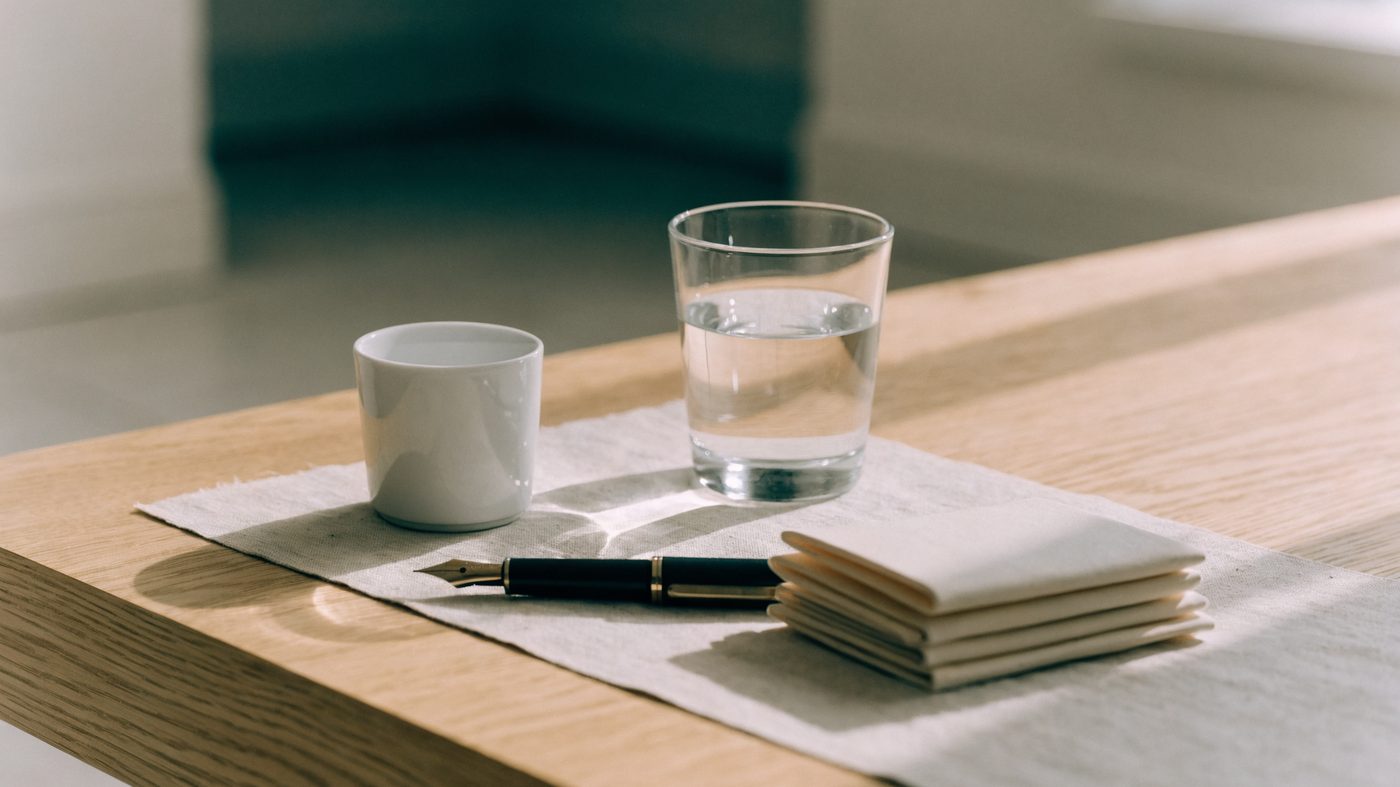 A pale oak desk with a fanned stack of folded cream cards, a fountain pen, a cup and a glass of water on a linen mat