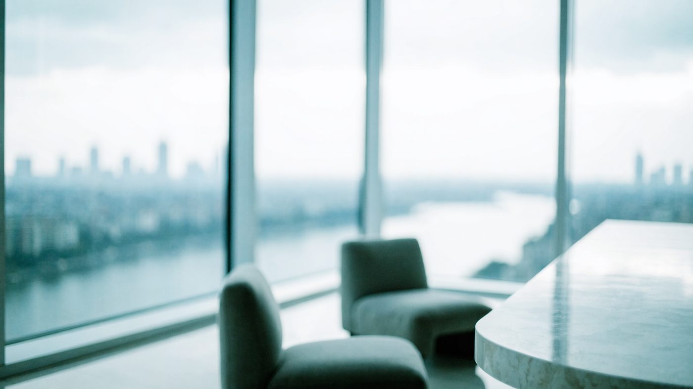 A high-floor room in a glass tower, pale stone table and low chairs against floor-to-ceiling glazing above the river