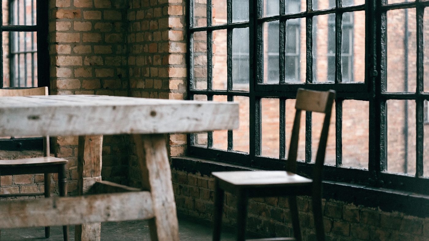 A converted warehouse room with exposed brick and black steel windows, a reclaimed timber table and two simple chairs