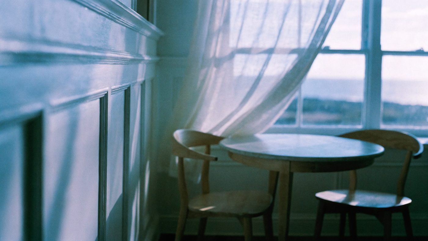 A bright seaside room with pale panelling, a small round table and two chairs beside a window filled with soft coastal light